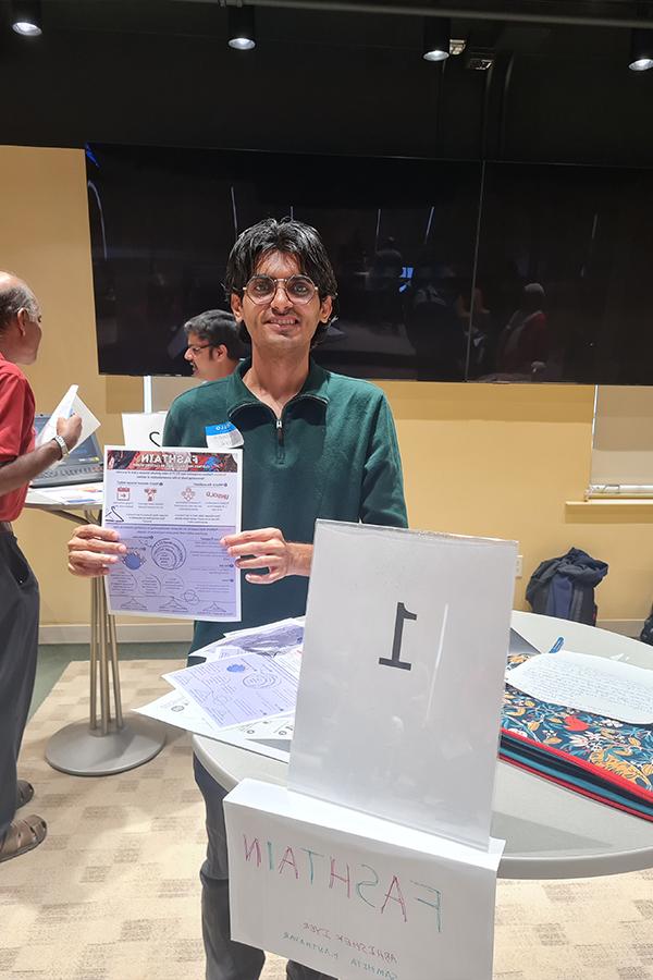 Abhishek is standing at a circular table holding a flyer with information about his startup, Fashtain